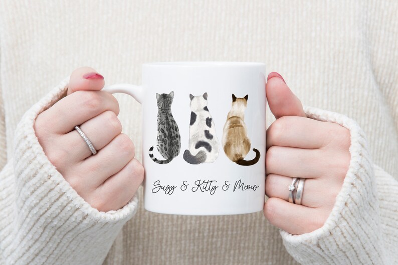 May include: White ceramic mug with three cats sitting back to back. The cats are a tabby, a black and white cat, and a Siamese. The mug has the text "Suzy & Kitty & Meow" printed on the bottom.