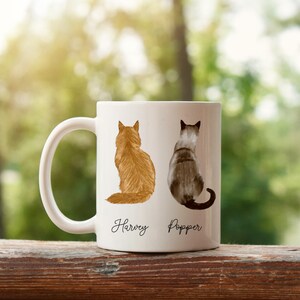 May include: White ceramic mug with a brown and orange cat on the left and a gray and white cat on the right. The cats are facing away from the viewer. The mug has the names "Harvey" and "Popper" written below the cats.