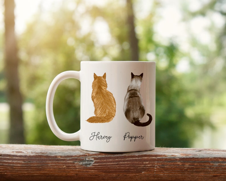 May include: White ceramic mug with a brown and orange cat on the left and a gray and white cat on the right. The cats are facing away from the viewer. The mug has the names "Harvey" and "Popper" written below the cats.