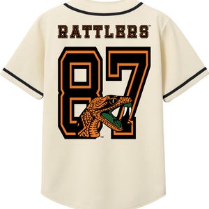 May include: Cream-colored baseball jersey with black trim and short sleeves. The back features the word "Rattlers" above the number "87" in black and orange. A snake graphic is incorporated into the number design.