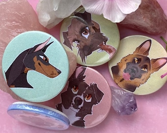 May include: Five round pins with different dog breeds on them. The pins are on a pink surface with a pink flower and a clear crystal.
