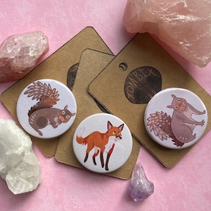 May include: Three round pinback buttons featuring illustrations of woodland animals. The buttons are white with a black outline. The first button shows a brown squirrel with a bushy tail. The second button shows a red fox with a white chest. The third button shows a gray squirrel with a bushy tail.