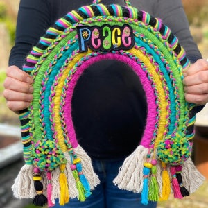 May include: A colourful rainbow-shaped wall hanging made of yarn with a peace sign in the centre. The rainbow is made of pink, yellow, green, blue, and black yarn. The peace sign is made of green felt with white stars and the word "peace" in black.