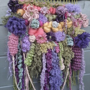 May include: A colorful wall hanging made of yarn in shades of pink, purple, green, and yellow. The yarn is woven into a variety of textures and patterns, creating a unique and eye-catching piece of art.