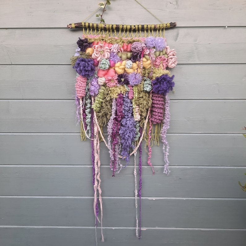 May include: A colorful wall hanging made of yarn and fabric. The hanging features a variety of textures and colors, including pink, purple, green, and yellow. The hanging is attached to a wooden dowel.