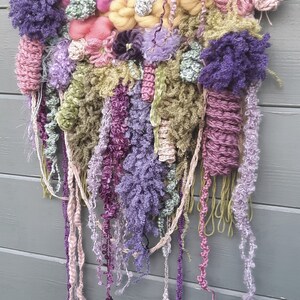 May include: A colorful wall hanging made of yarn and fabric in shades of pink, purple, green, and yellow. The hanging is woven and has a variety of textures, including pom-poms, fringe, and tassels.
