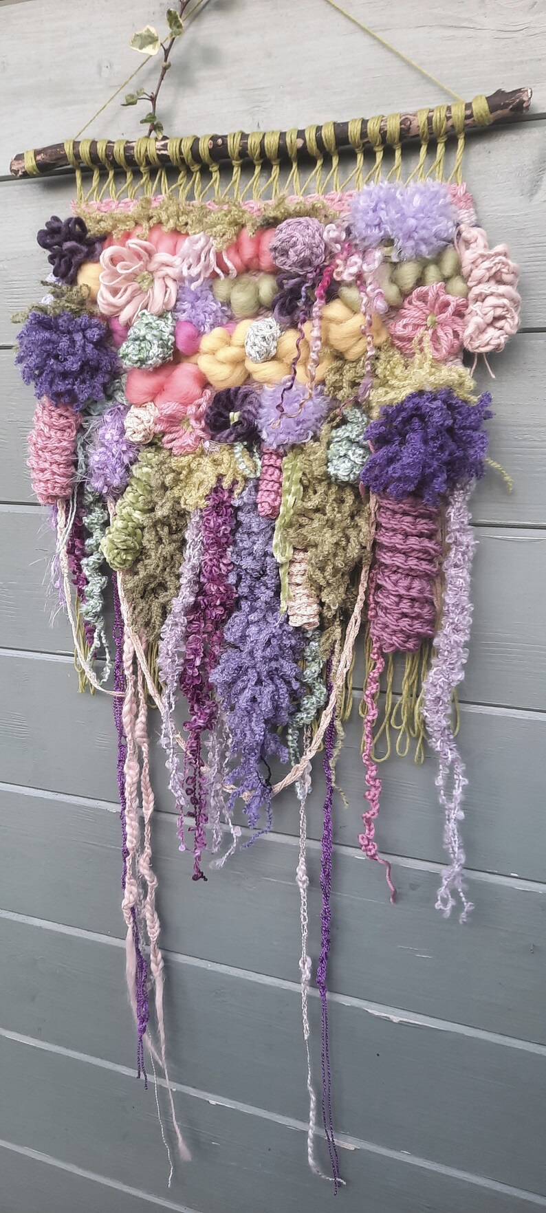 May include: A colorful wall hanging made of yarn and fabric in shades of pink, purple, green, and yellow. The hanging is woven and has a variety of textures, including pom-poms, fringe, and tassels.