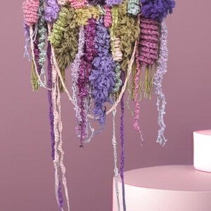 May include: A colorful wall hanging made of yarn and fabric. The hanging features a variety of textures and colors, including pink, purple, green, and yellow. The hanging is attached to a wooden dowel.