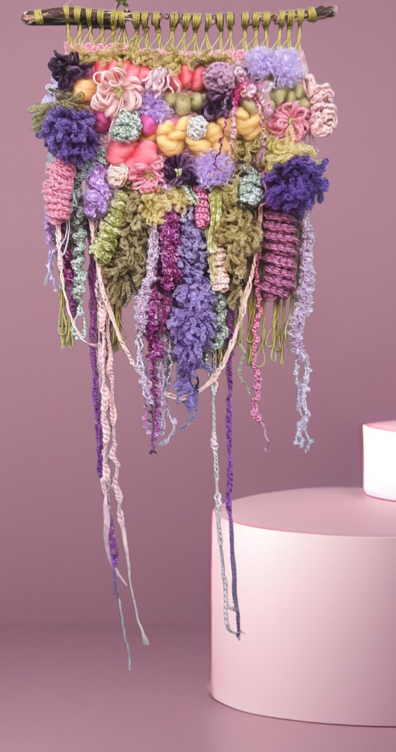 May include: A colorful wall hanging made of yarn and fabric. The hanging features a variety of textures and colors, including pink, purple, green, and yellow. The hanging is attached to a wooden dowel.