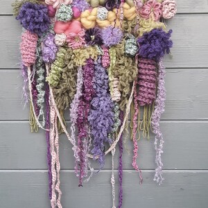 May include: A colorful wall hanging made of yarn and fabric. The hanging features a variety of textures and colors, including pink, purple, green, and yellow. It is woven on a wooden stick and has long strands of yarn hanging down.