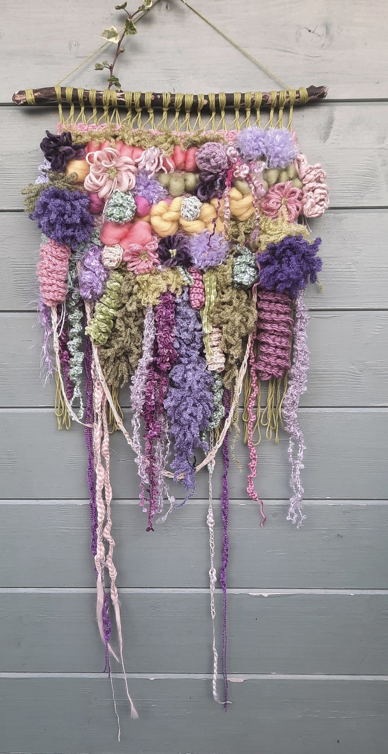 May include: A colorful wall hanging made of yarn and fabric. The hanging features a variety of textures and colors, including pink, purple, green, and yellow. It is woven on a wooden stick and has long strands of yarn hanging down.