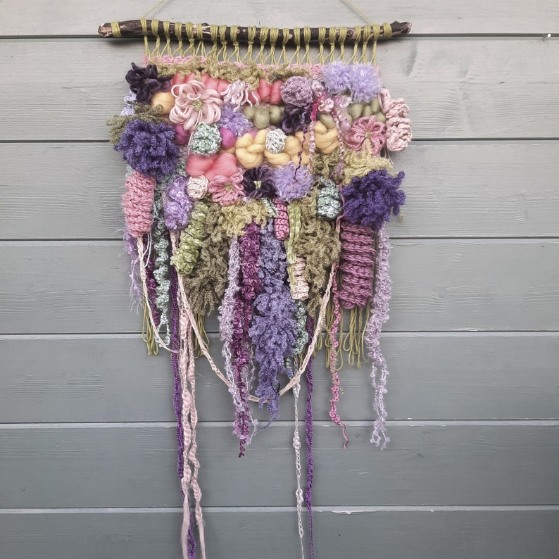 May include: A colorful wall hanging made of yarn and fabric in shades of pink, purple, and green. The hanging is woven and has a fringe at the bottom.