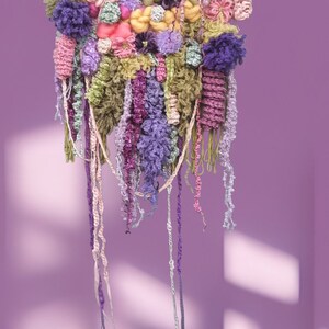 May include: A colorful wall hanging made of yarn and fabric. The hanging features a variety of textures and colors, including pink, purple, green, and yellow. The hanging is attached to a wooden frame and has long strands of yarn hanging down from the bottom.
