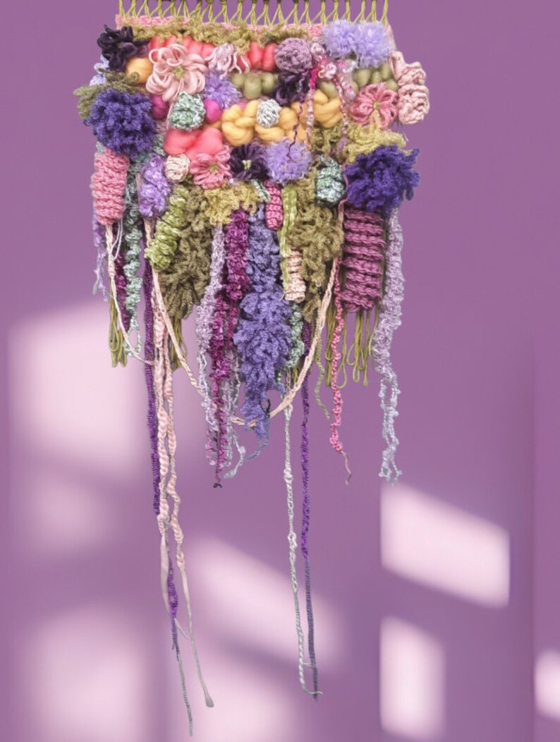 May include: A colorful wall hanging made of yarn and fabric. The hanging features a variety of textures and colors, including pink, purple, green, and yellow. The hanging is attached to a wooden frame and has long strands of yarn hanging down from the bottom.