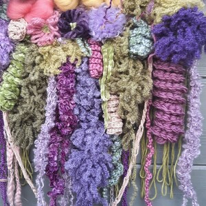 May include: A colorful wall hanging made of yarn and fabric. The hanging features a variety of textures and colors, including pink, purple, green, and yellow. The hanging is attached to a wooden dowel.