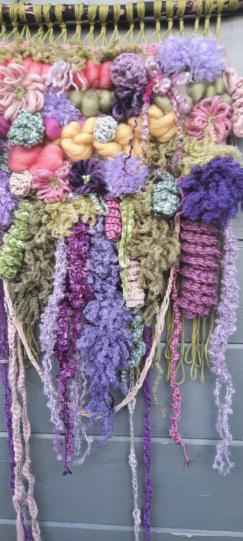May include: A colorful wall hanging made of yarn and fabric. The hanging features a variety of textures and colors, including pink, purple, green, and yellow. The hanging is attached to a wooden dowel.