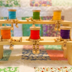 May include: Six wooden spools of thread with articulated wooden limbs, each wrapped in a different color of thread including blue, green, yellow, orange, red, and purple. The spools are arranged sitting on two tiered light-colored wooden shelves.