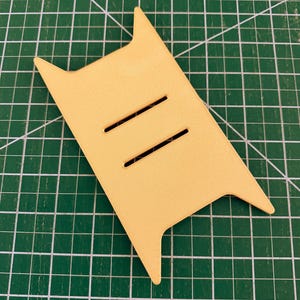May include: A light yellow leather piece with two parallel slits and pointed edges. The shape is rectangular with two extended points at the top and two at the bottom. The item is on a green cutting mat.