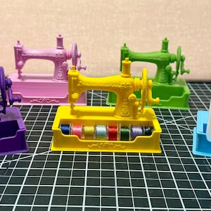 May include: A collection of seven plastic sewing machine-shaped business card holders in red, purple, pink, yellow, green, orange, and blue. The yellow holder contains small thread spools, and the blue holder displays a business card for Yellow Rose Quilts.
