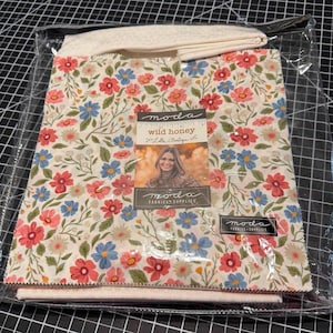 May include: A stack of fabric squares in a clear plastic package, featuring a floral pattern with red, blue, and green flowers on a cream background. A label on the front reads "moda wild honey" with a photograph of a person.