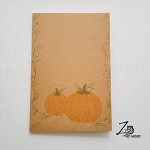 May include: A brown paper notebook with a green vine border and two orange pumpkins drawn on the front. The pumpkins have a detailed, intricate design.