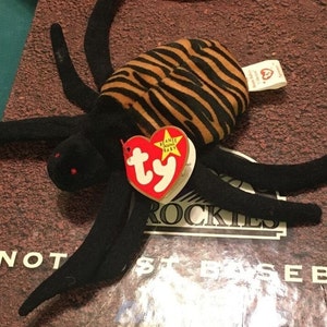 Può includere: Ragno di peluche Beanie Baby a strisce nere e arancioni con un'etichetta Ty rossa che dice "Beanie Babies" e una stella. L'etichetta è attaccata alla schiena del ragno.