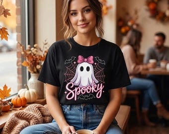 May include: A black short-sleeved t-shirt featuring a graphic of a white ghost with a pink bow, surrounded by spider webs, stars, and a bat. The word Spooky is printed in a neon pink script font below the ghost illustration on the front.