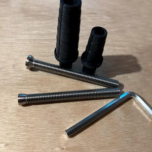 以下が含まれることがあります： 2つの黒いゴムブッシュ、2つの銀色のねじ付きボルト、銀色の六角レンチを含むハードウェアコンポーネントの品揃え。アイテムは、明るい茶色の木製の表面に配置されています。