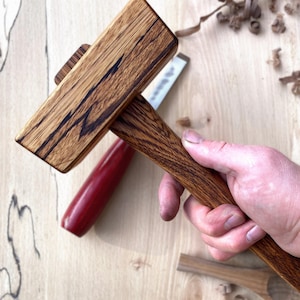 May include: A handcrafted wooden mallet featuring a rectangular head and a rounded handle. The mallet is made from light brown wood with dark brown grain. A red-handled chisel and other woodworking tools are visible in the background.