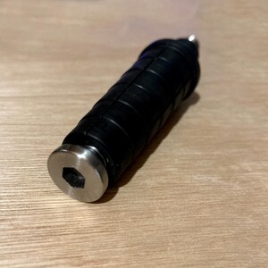以下が含まれることがあります： 黒色の円筒形の物体で、テクスチャ加工されたグリップと銀色の金属製のエンドキャップが付いています。エンドキャップには六角形のくぼみがあります。明るい茶色の木製の表面に置かれています。