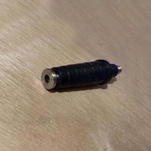 以下が含まれることがあります： 黒い円筒形の物体で、溝付きのゴム製グリップと銀色の金属製の端が付いています。端は六角形です。物体は明るい茶色の木製の表面に置かれています。