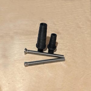 以下が含まれることがあります： 2つの黒いゴム製グロメットと2つの銀色の金属ネジが、明るい茶色の木製の表面に配置されています。グロメットは、リブ付きのテクスチャを持つ円筒形です。ネジはねじ山のあるデザインと平らな頭を持っています。