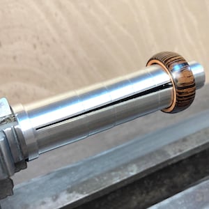 May include: A wooden ring with a striped pattern is mounted on a silver metal tool. The ring has a dark brown and tan color scheme. The tool appears to be part of a larger machine, possibly for woodworking or jewelry making.