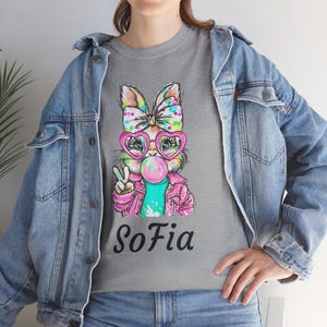 May include: A gray t-shirt featuring a cartoon rabbit wearing heart-shaped glasses and blowing a bubblegum bubble. The rabbit is wearing a pink jacket and a bow. The name "Sofia" is printed below the rabbit.