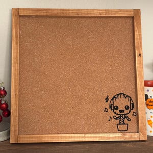 以下が含まれることがあります： 木製の枠が付いた茶色のコルクボード。コルクボードには、ヘッドホンを付けた漫画のキャラクターの白黒のイラストが描かれています。