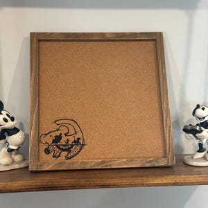May include: A square corkboard with a dark wooden frame. A black graphic of a lion cub is in the lower left corner. The corkboard is displayed on a dark wooden shelf, flanked by Mickey Mouse figurines.