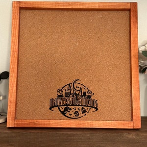 May include: A brown wooden framed corkboard with a black Universal Studios logo featuring various attractions including the Hogwarts Express, Jurassic Park, and the Minions.