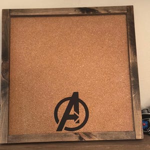 May include: A brown corkboard with a wooden frame. The corkboard has a black vinyl sticker of the Avengers logo on it.