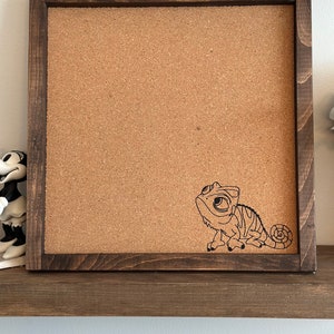 May include: A brown wooden framed corkboard with a black line drawing of a cartoon chameleon on the cork surface.