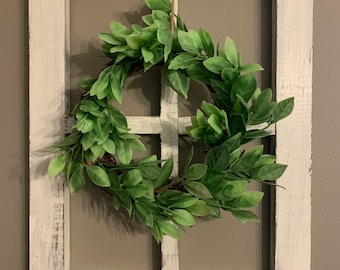 May include: A white wooden window frame with a green leafy wreath hanging from the top. The wreath is made of artificial greenery and is hanging in the center of the frame.