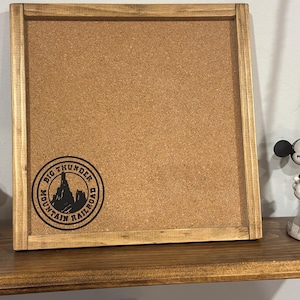 Puede incluir: Un corcho enmarcado en madera con las palabras "Big Thunder Mountain Railroad" en un diseño circular. El corcho es de color marrón claro y el marco es de color marrón más oscuro. El diseño es negro.