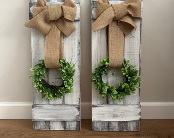 May include: Two white wooden shutters with a burlap bow and a green wreath on each. The shutters are distressed and have a rustic look.