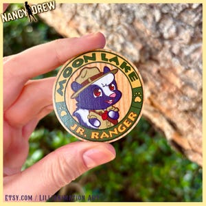 May include: A round wooden Moon Lake Jr. Ranger badge featuring a cartoon skunk in a ranger hat and uniform. The badge has green and gold lettering and a paw print design. The Nancy Drew logo is in the top left corner.