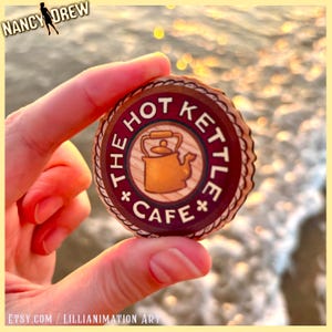 Puede incluir: Un pin redondo de madera con temática de Nancy Drew que presenta "The Hot Kettle Cafe" en texto blanco alrededor de una imagen central de una tetera dorada. El pin se sostiene entre los dedos contra un fondo borroso del océano.
