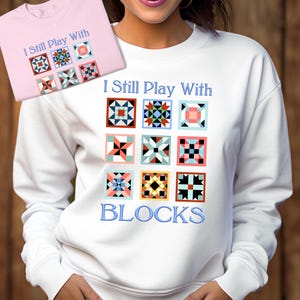 May include: White sweatshirt with the text "I Still Play With BLOCKS" in blue. The sweatshirt features a design of nine colorful quilt blocks.
