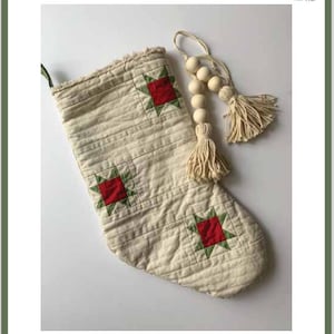 Pode incluir: Uma meia de Natal acolchoada branca com remendos em forma de estrela vermelha e verde. A meia tem uma borda desfiada e é decorada com uma guirlanda de contas de madeira e um tassel branco.