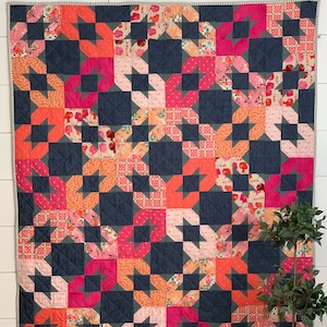 Puede incluir: Una colcha con fondo azul marino y un patrón de estrellas rosas, naranjas y blancas. La colcha está hecha con una variedad de telas y tiene un diseño simple y moderno.