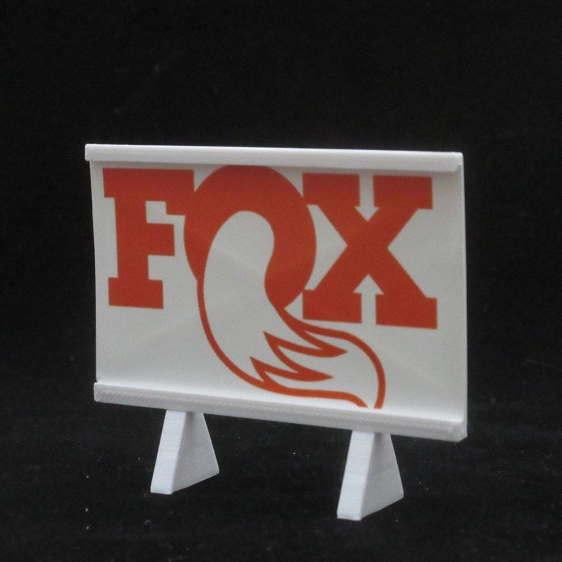 May include: White display stand with a white sign featuring the word "FOX" in red letters and a red fox tail logo.