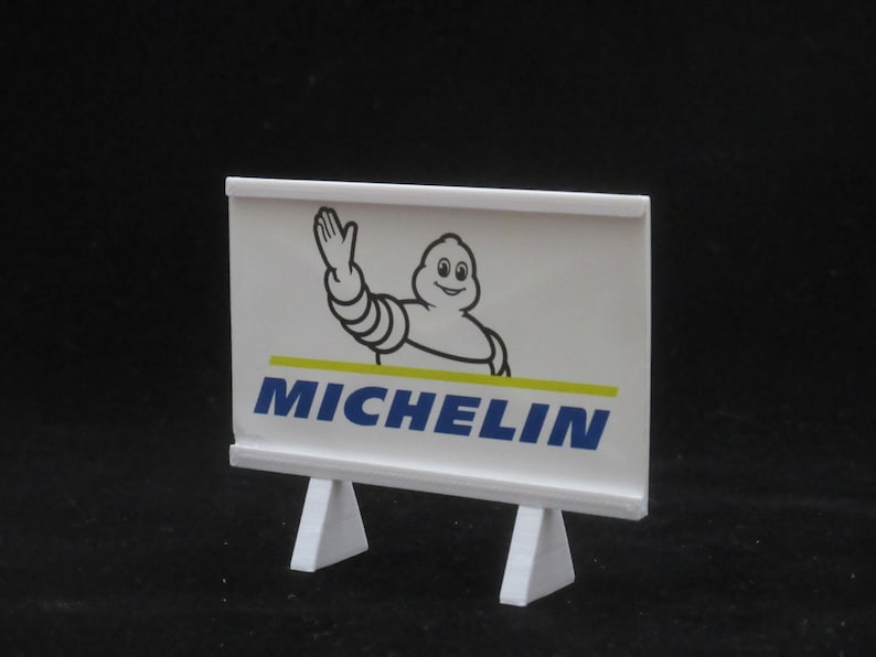 May include: White sign with a cartoon Michelin Man waving and the word "MICHELIN" in blue letters. The sign is on a white stand with two triangular legs.