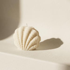 Könnte beinhalten: Eine cremefarbene Wachskerze in Form einer abgerundeten Muschel mit einer texturierten, gerippten Oberfläche. Die Kerze hat einen einzelnen, mittig platzierten Docht an der Oberseite und wirft einen deutlichen Schatten auf die rechte Seite.